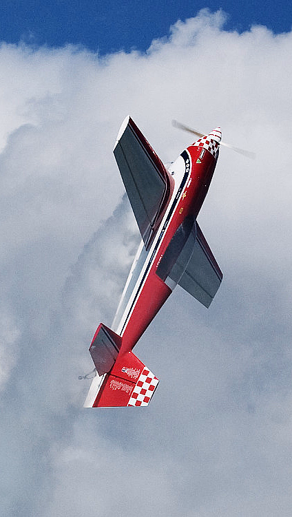 Et rødt akrobatfly med hvite striper som flyr vertikalt mot en blå himmel med hvite skyer.