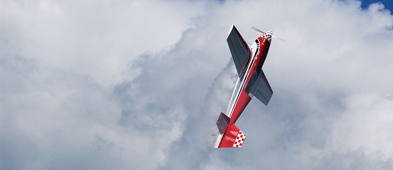 Et rødt akrobatfly med hvite striper som flyr vertikalt mot en blå himmel med hvite skyer.