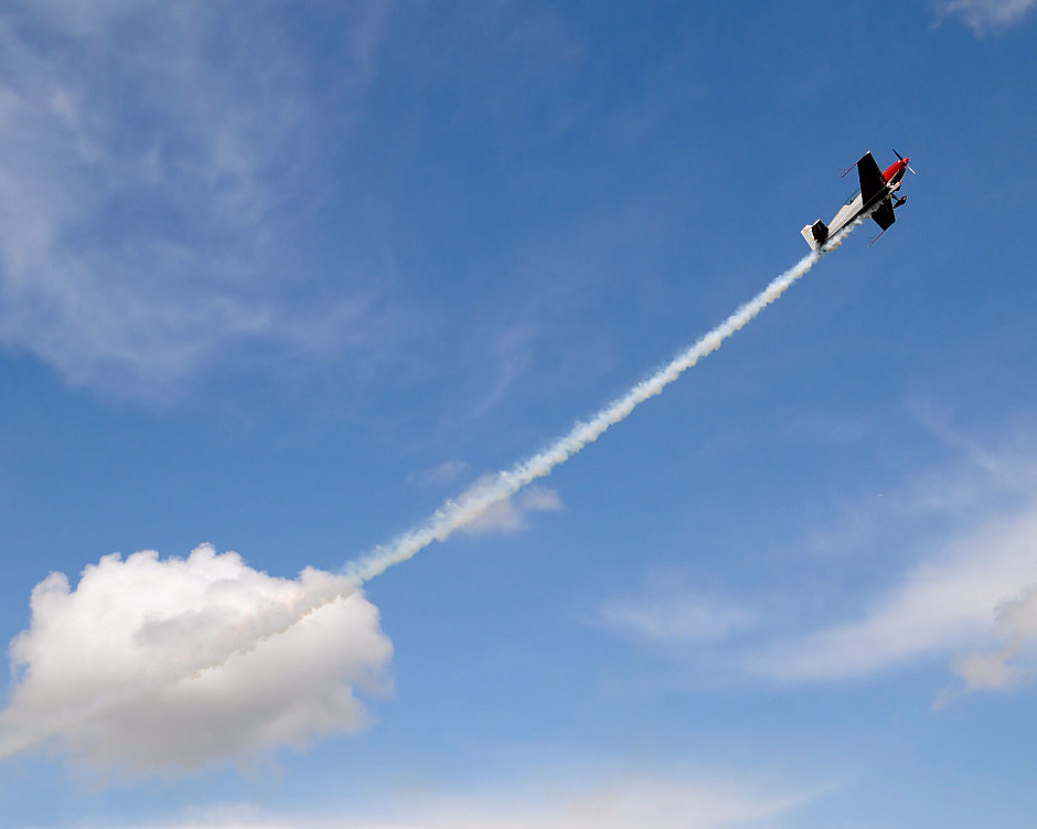 Et lite fly med rød nese og svart kropp flyr oppover i en blå himmel og etterlater seg en hvit røykstripe blant hvite skyer.