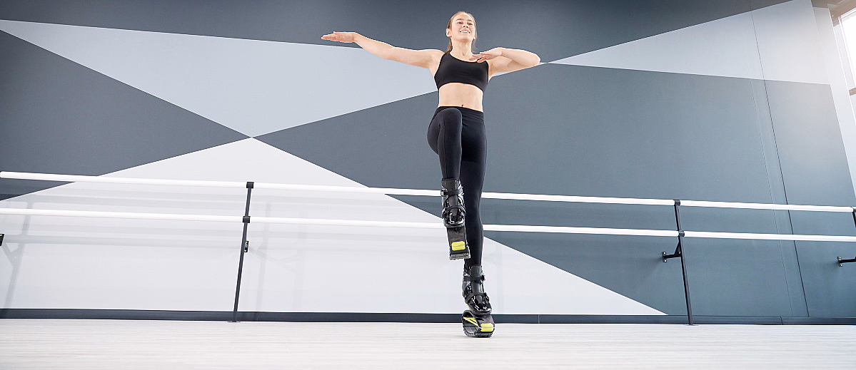 En person i svarte treningsklær balanserer på ett ben med Kangoo Jump-støvler i et moderne studio med geometrisk veggmønster.