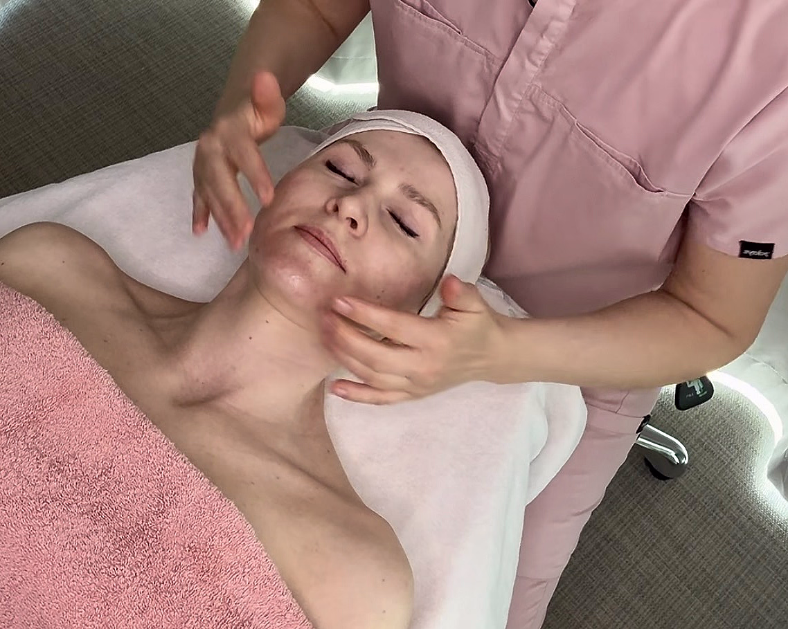 En person får en avslappende ansiktsmassasje av en behandler på et spa, liggende på en behandlingsbenk med et rosa håndkle.