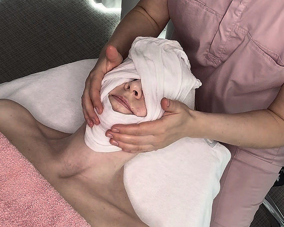 En person som ligger ned under en spabehandling med et hvitt håndkle pakket rundt hodet og øynene, og et rosa håndkle over brystet.