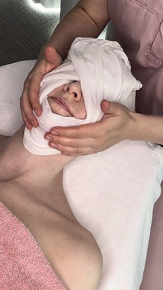 En person som ligger ned under en spabehandling med et hvitt håndkle pakket rundt hodet og øynene, og et rosa håndkle over brystet.