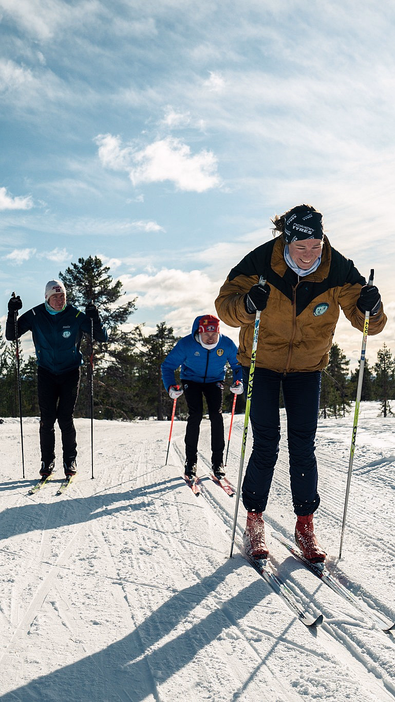 Tre personer går på langrenn i en snødekt løype under en blå himmel. Den fremste skiløperen har på seg en brun jakke og svarte bukser, mens de andre følger etter i blå jakker med grantrær i bakgrunnen.