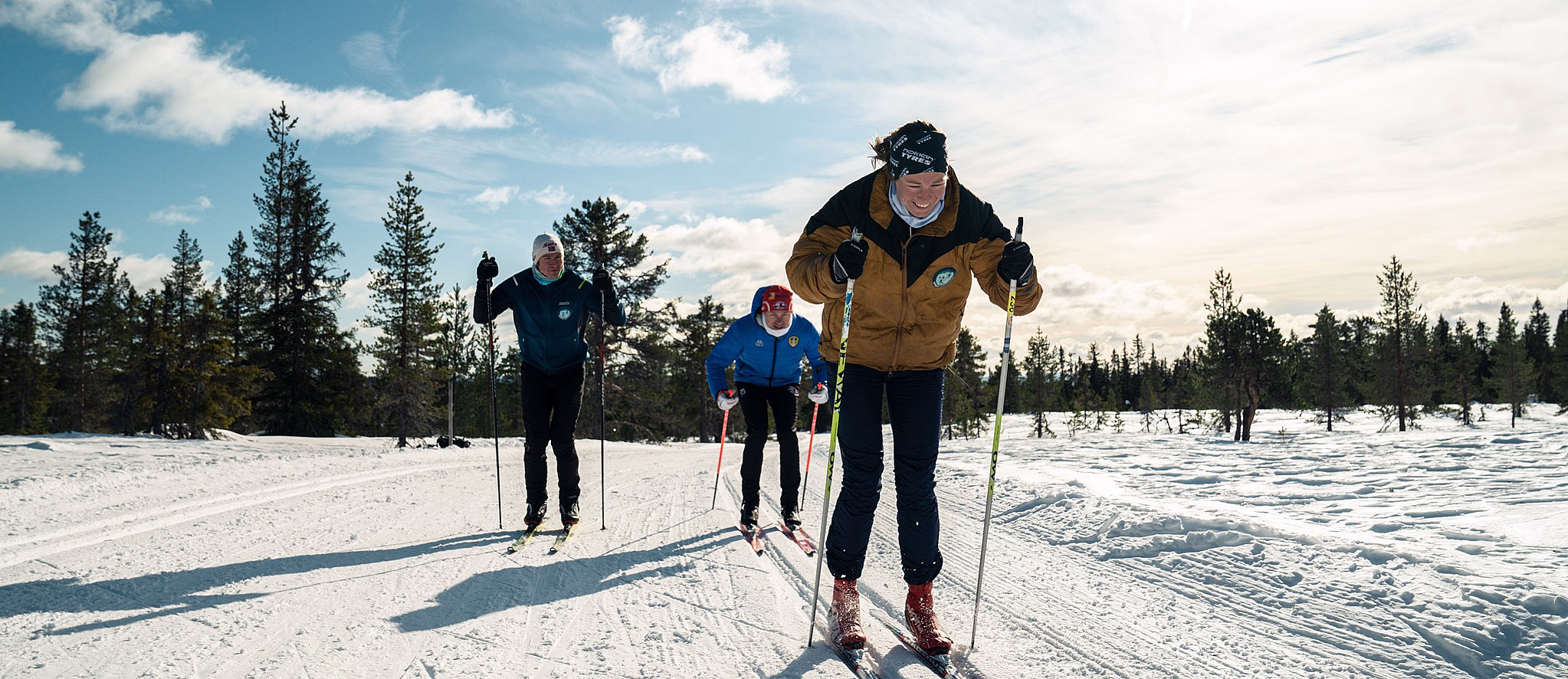 Tre personer går på langrenn i en snødekt løype under en blå himmel. Den fremste skiløperen har på seg en brun jakke og svarte bukser, mens de andre følger etter i blå jakker med grantrær i bakgrunnen.
