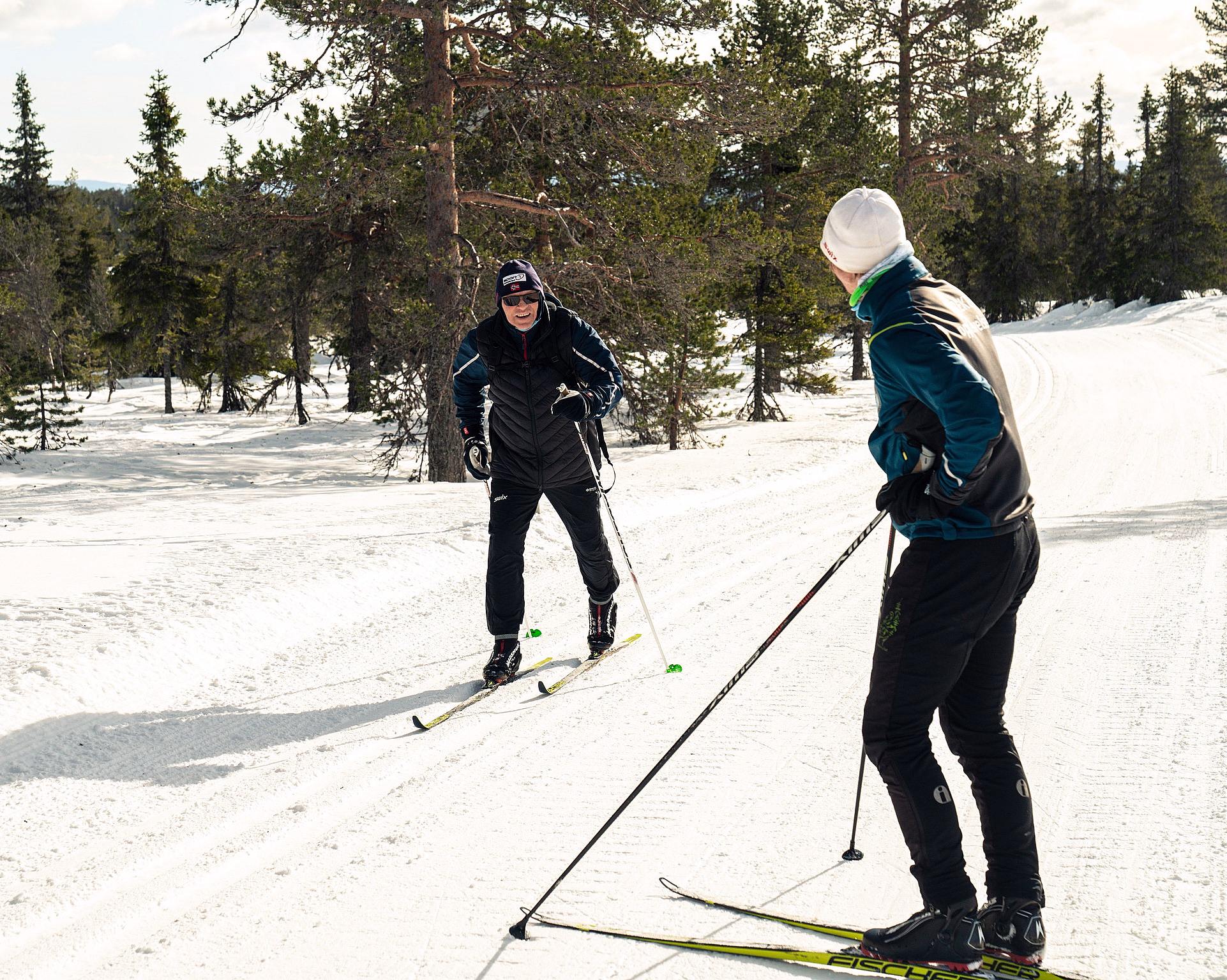 To personer som går på langrenn på en snødekt sti omgitt av høye trær.