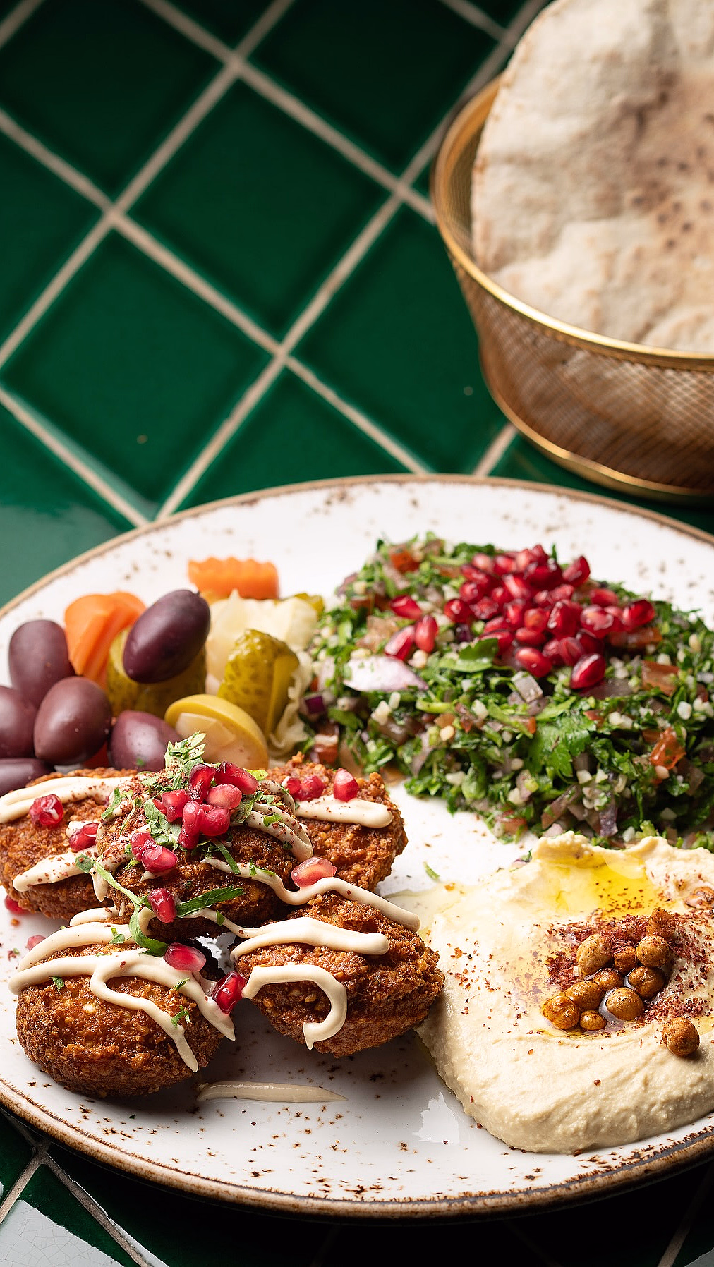 Hvit tallerken med falafel, hummus, tabbouleh, syltede grønnsaker og oliven, servert med pitabrød på en grønn flisoverflate.