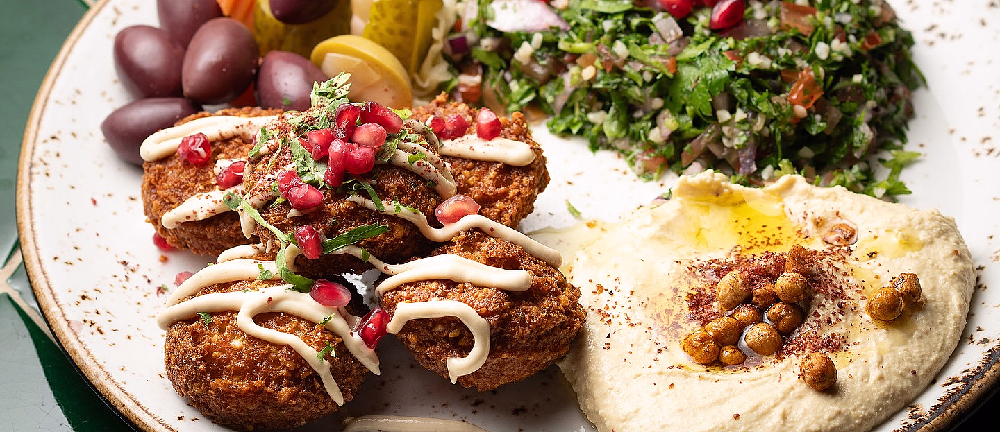 Hvit tallerken med falafel, hummus, tabbouleh, syltede grønnsaker og oliven, servert med pitabrød på en grønn flisoverflate.