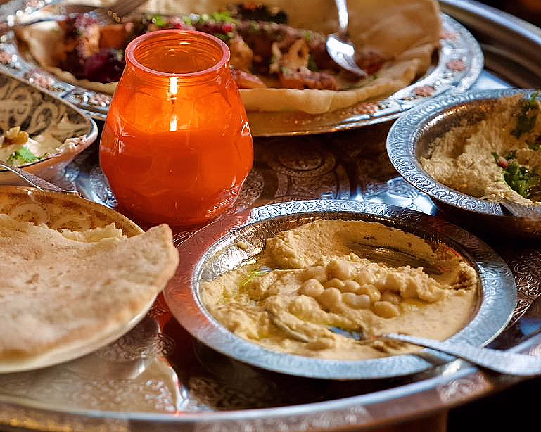Et rundt metallbord med intrikate mønstre, dekket med tallerkener med hummus, pitabrød og grillet kjøtt, samt et brennende oransje lys.