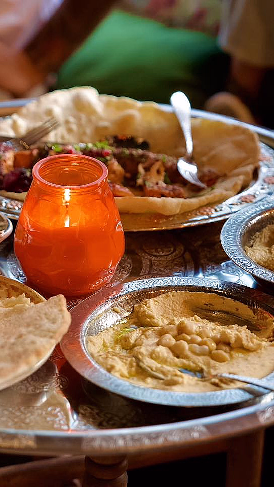 Et rundt metallbord med intrikate mønstre, dekket med tallerkener med hummus, pitabrød og grillet kjøtt, samt et brennende oransje lys.