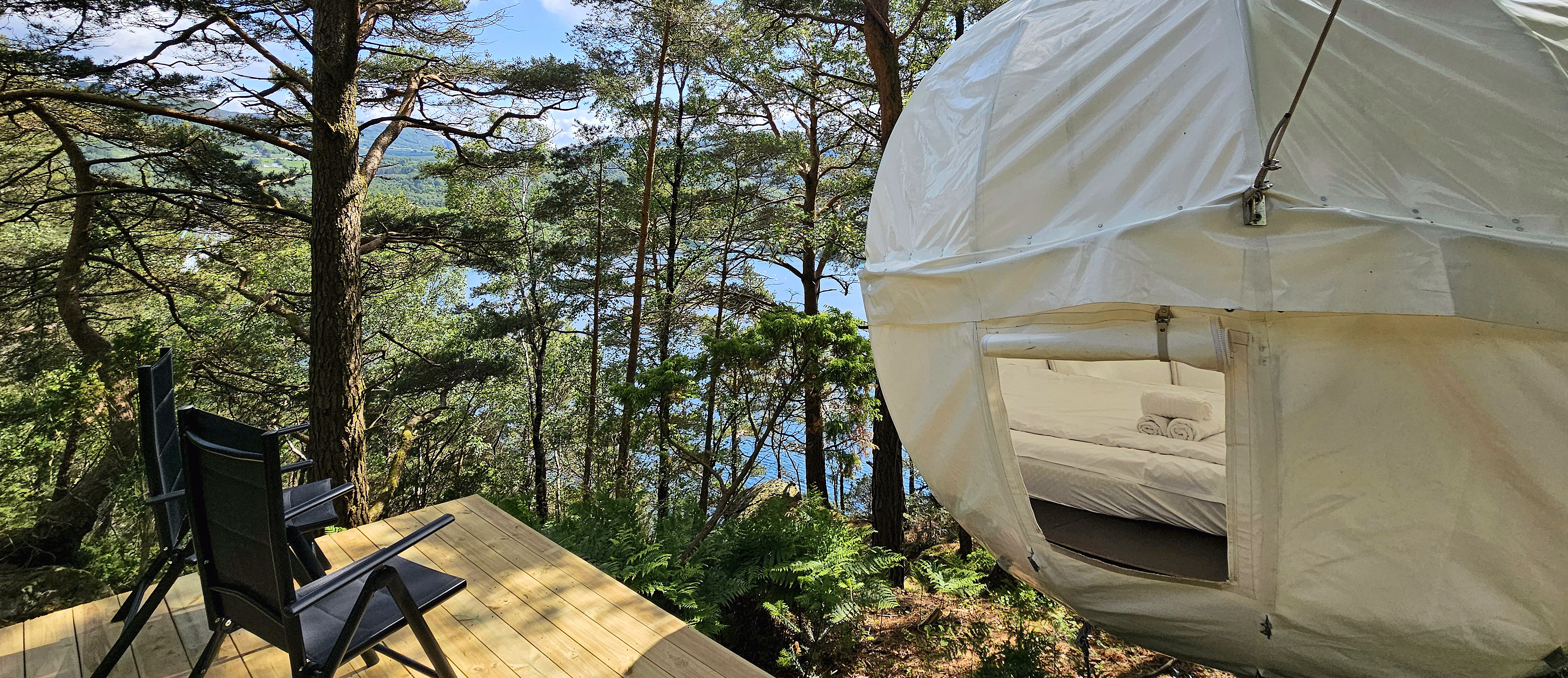 Et hvitt, kuleformet telt henger mellom høye furutrær over en treterrasse med to svarte stoler i et solfylt skoglandskap.