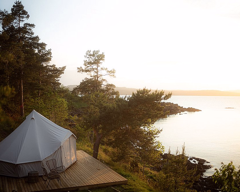 Et hvitt glampingtelt på en treterrasse ved et stille vann, omgitt av grønne trær med to stoler og et bord på dekket.