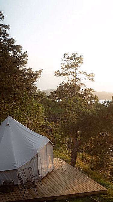 Et hvitt glampingtelt på en treterrasse ved et stille vann, omgitt av grønne trær med to stoler og et bord på dekket.