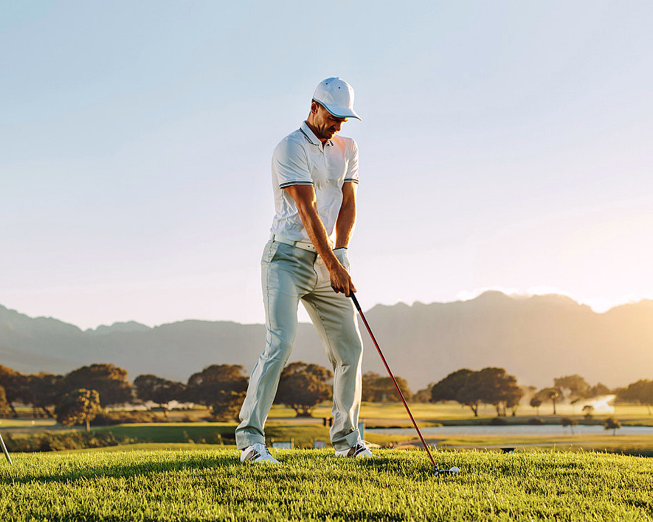 En golfspiller i hvit skjorte og caps forbereder seg på å slå en golfball på en grønn bane med fjell i bakgrunnen.