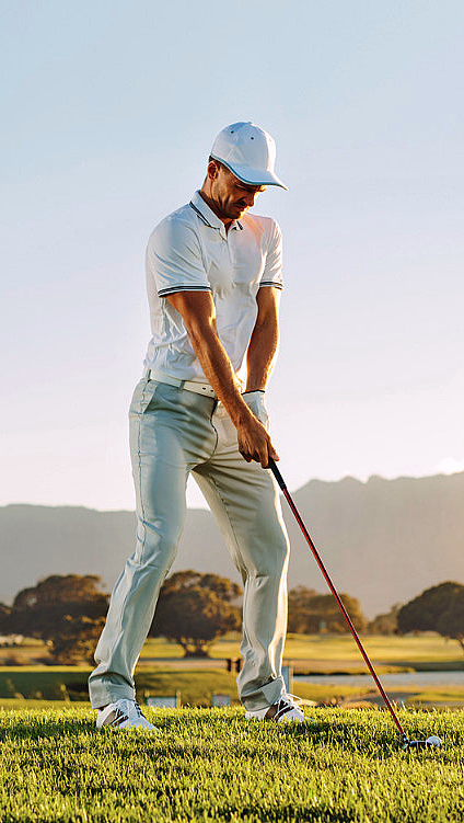 En golfspiller i hvit skjorte og caps forbereder seg på å slå en golfball på en grønn bane med fjell i bakgrunnen.