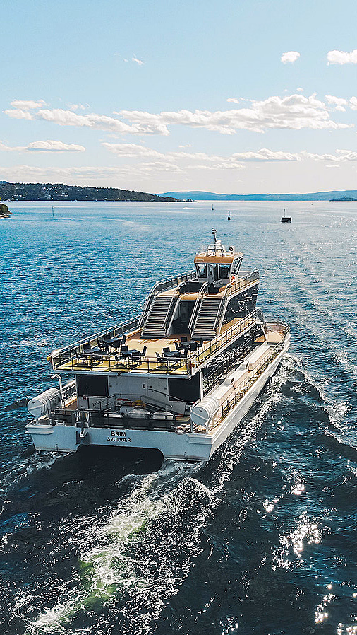 En hvit ferge seiler på et glitrende blått hav med grønne åser i bakgrunnen.
