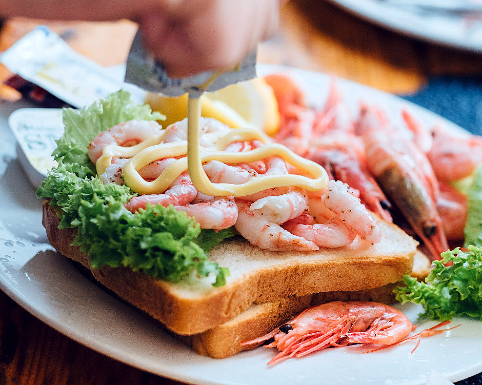 En hånd spruter majones på et rekesmørbrød med ferske reker og grønn salat på en hvit tallerken.