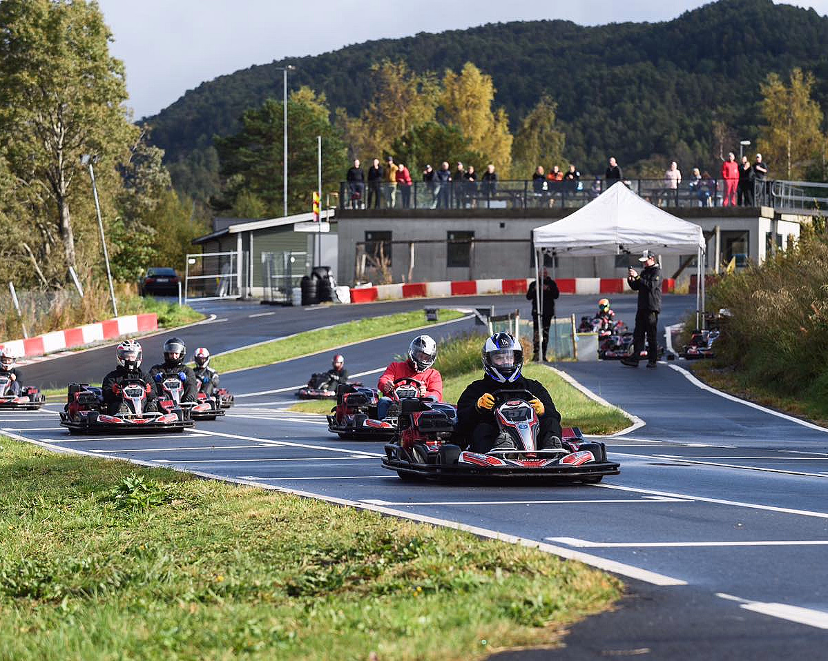 Flere personer kjører gokart på en utendørsbane med tilskuere i bakgrunnen