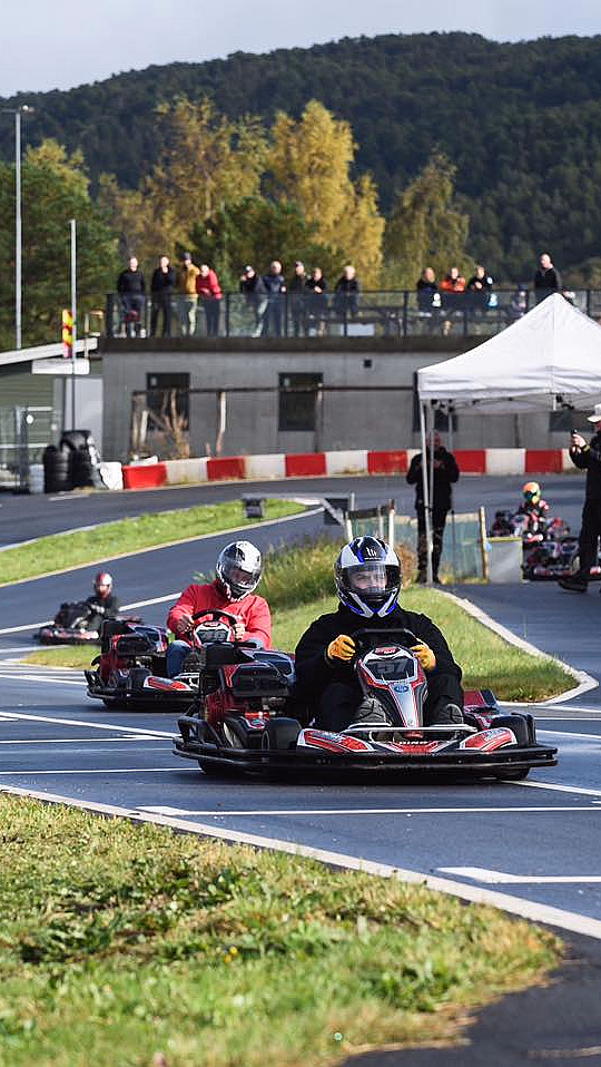 Flere personer kjører gokart på en utendørsbane med tilskuere i bakgrunnen