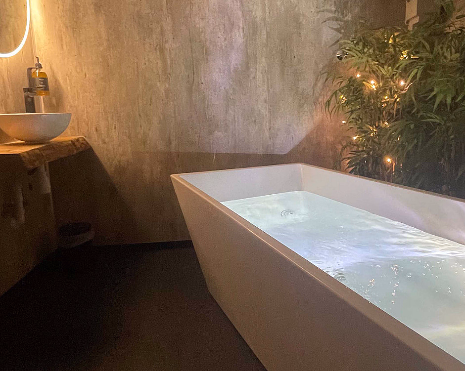 Moderne baderom med et hvitt rektangulært badekar, et rundt speil med baklys over en servant i tre, og en grønn plante med små lyslenker.