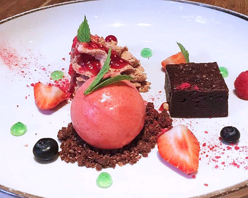 Desserttallerken med en kule rosa sorbet, sjokoladebrownie, friske bær og dekorative sauser på et trebord.