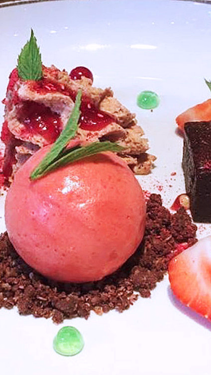 Desserttallerken med en kule rosa sorbet, sjokoladebrownie, friske bær og dekorative sauser på et trebord.