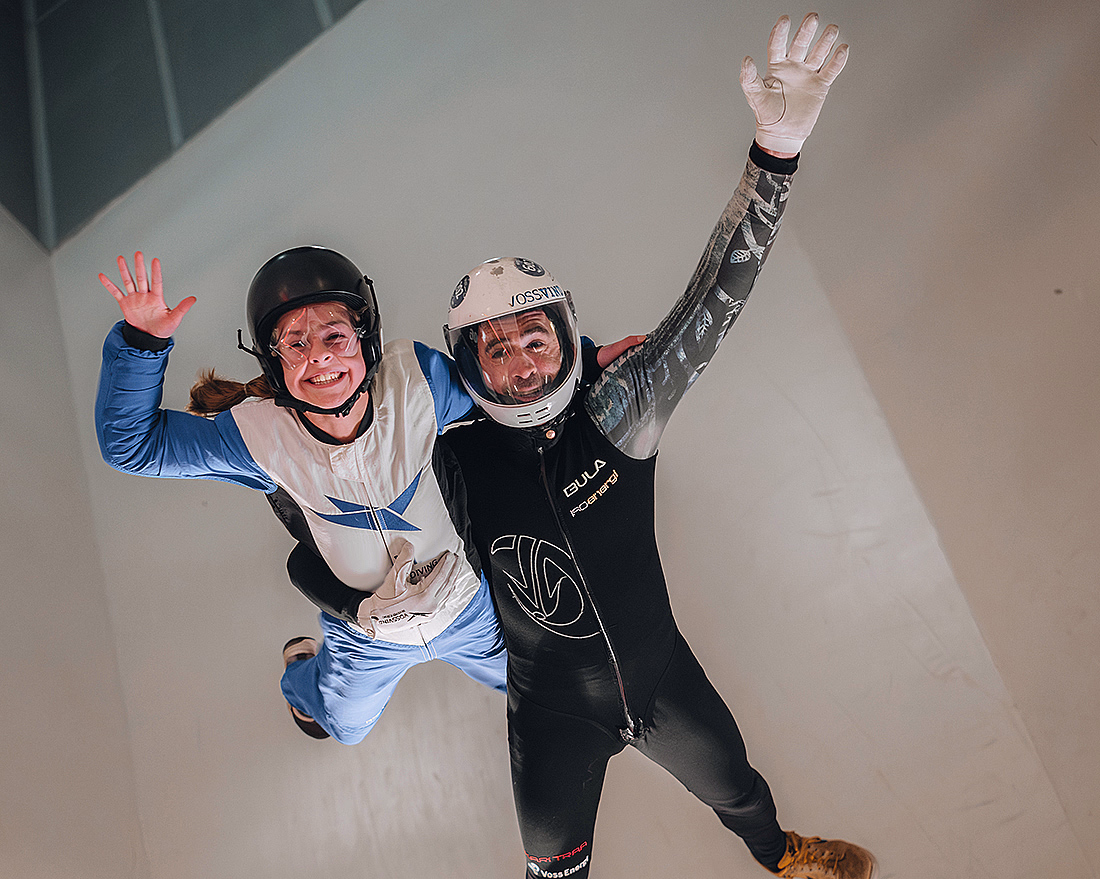 To personer i flyvedrakter og hjelmer som smiler og vinker mens de svever i en vindtunnel.