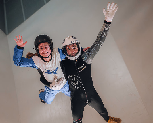 To personer i flyvedrakter og hjelmer som smiler og vinker mens de svever i en vindtunnel.