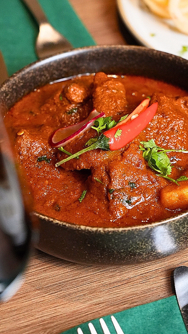 En mørk bolle med rød curry og kjøtt, toppet med chili og koriander, servert på et trebord med en grønn serviett.