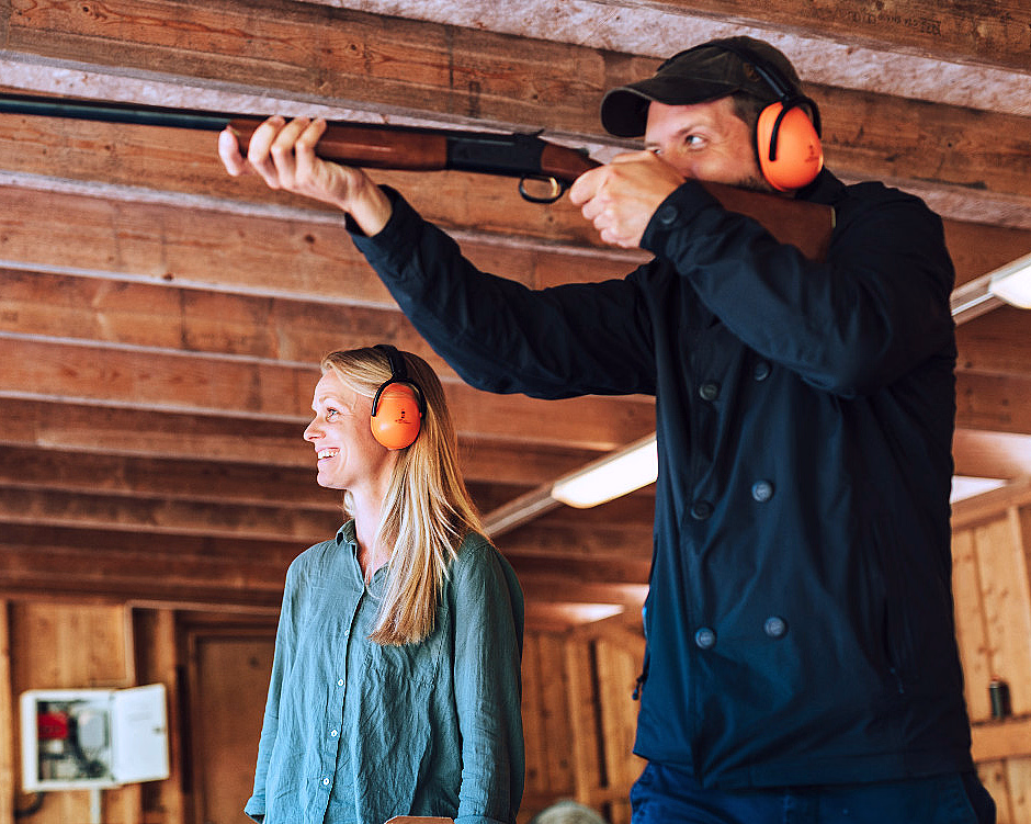 En mann sikter med en rifle på en innendørs skytebane i tre, mens en kvinne smiler ved siden av ham. Begge har på seg oransje hørselvern.