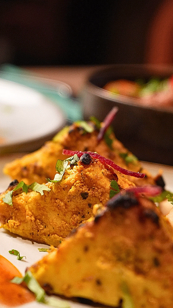 Nærbilde av gylne, trekantede stykker grillet paneer på en hvit tallerken, pyntet med grønne urter, rødløk og en oransje saus.