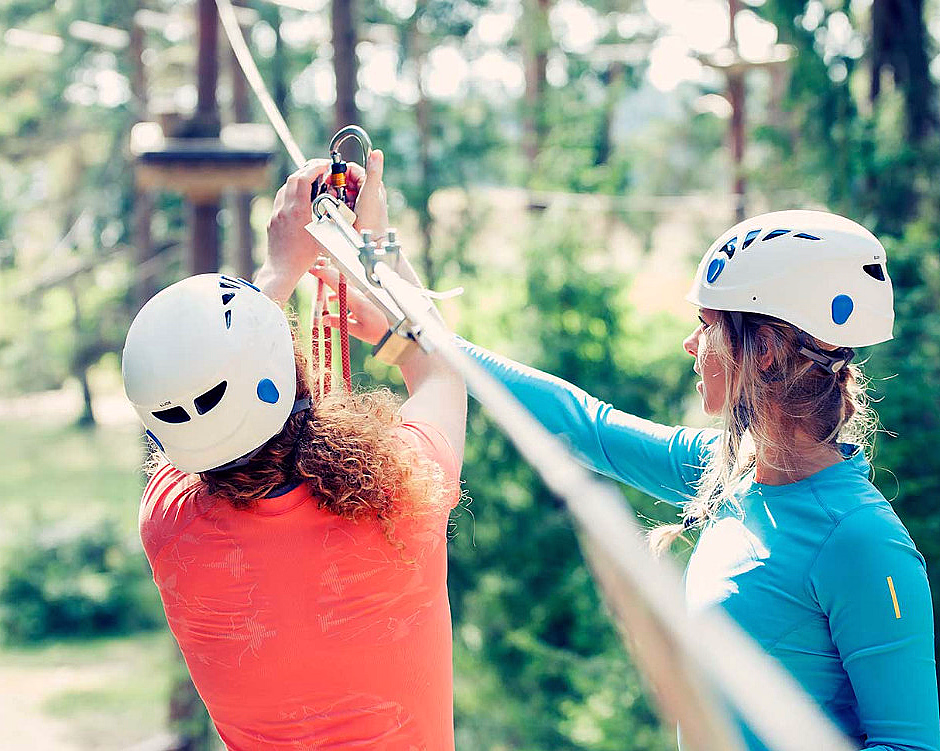 To personer med hvite hjelmer og klatreutstyr på en zipline i en frodig skog med sollys.