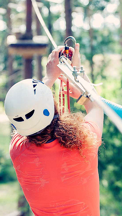 To personer med hvite hjelmer og klatreutstyr på en zipline i en frodig skog med sollys.