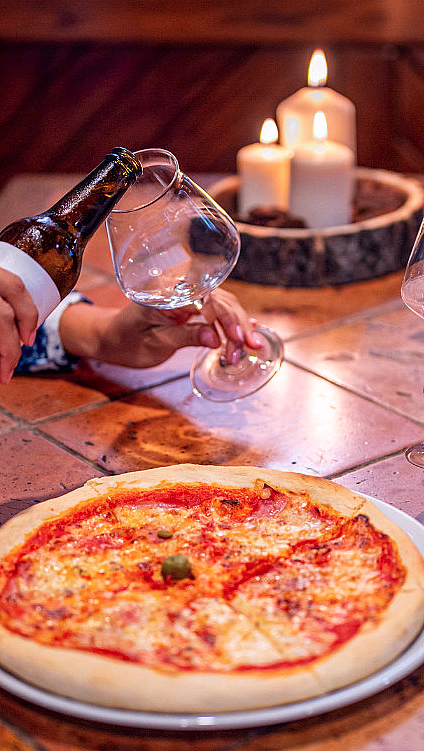 En pizza på et brunt flisbord med to personer som skjenker drikke i glass og tre tente stearinlys i bakgrunnen.