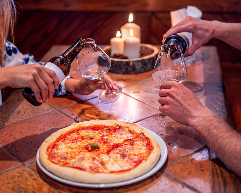 En pizza på et brunt flisbord med to personer som skjenker drikke i glass og tre tente stearinlys i bakgrunnen.