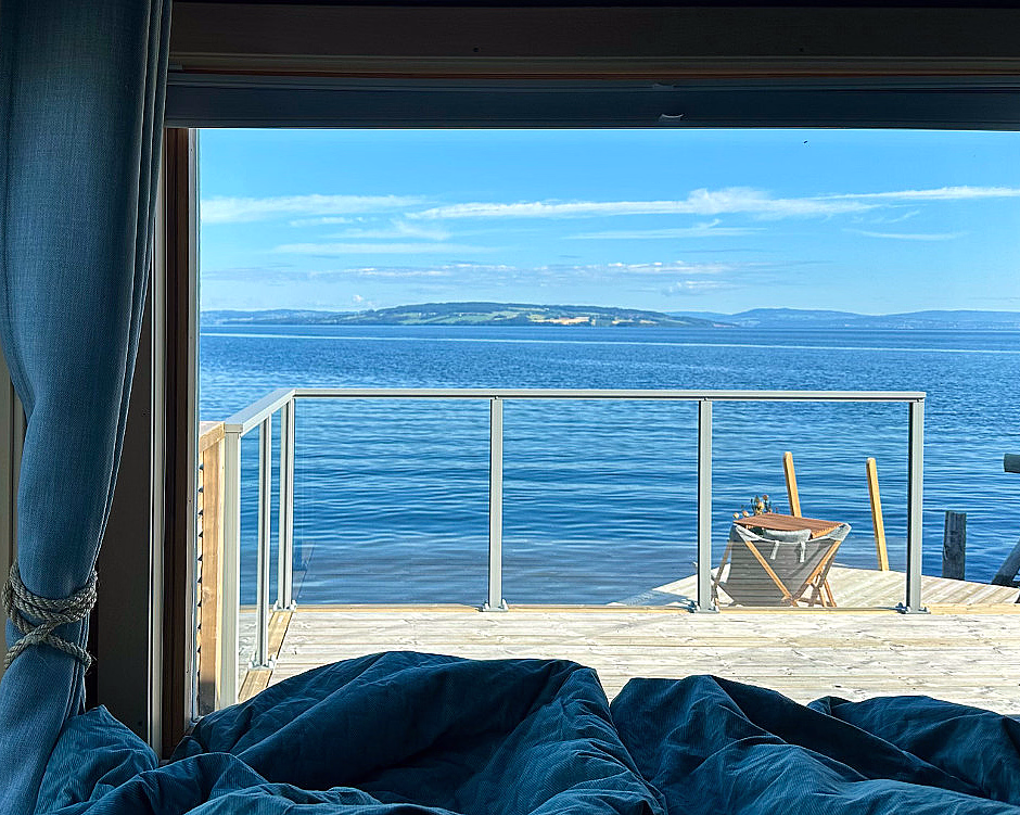 Utsikt fra et vindu med blå gardiner som rammer inn en terrasse med utemøbler, med utsikt over et blått vann og fjerne åser under en klar himmel.