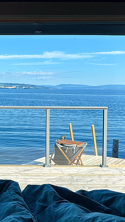 Utsikt fra et vindu med blå gardiner som rammer inn en terrasse med utemøbler, med utsikt over et blått vann og fjerne åser under en klar himmel.