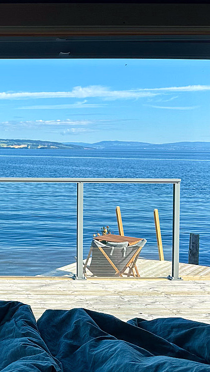 Utsikt fra et vindu med blå gardiner som rammer inn en terrasse med utemøbler, med utsikt over et blått vann og fjerne åser under en klar himmel.