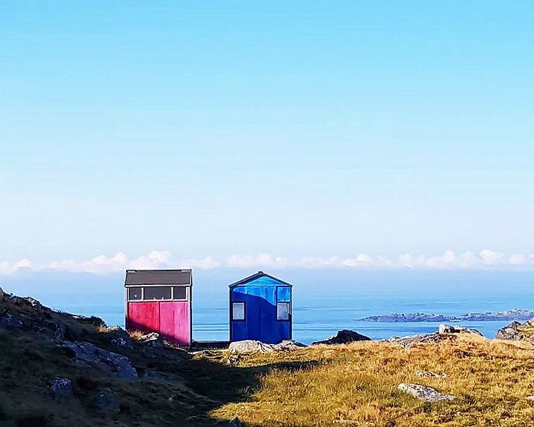 To små hytter, en rosa og en blå, på en gresskledd og steinete bakke ved havet under en blå himmel.