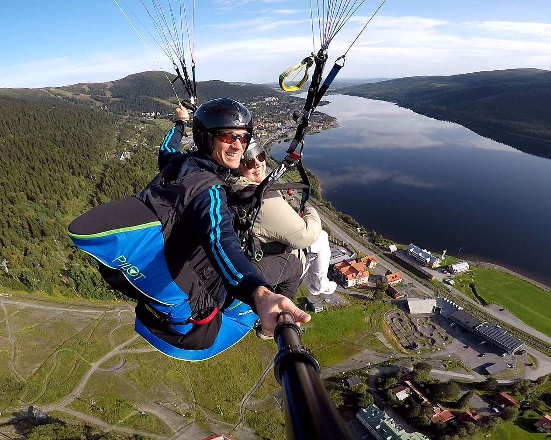 To personer som paraglider over et vidstrakt landskap med grønn skog, en blå innsjø og bebyggelse under en klar himmel.