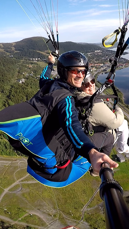 To personer som paraglider over et vidstrakt landskap med grønn skog, en blå innsjø og bebyggelse under en klar himmel.