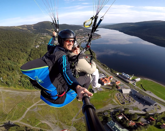 To personer som paraglider over et vidstrakt landskap med grønn skog, en blå innsjø og bebyggelse under en klar himmel.