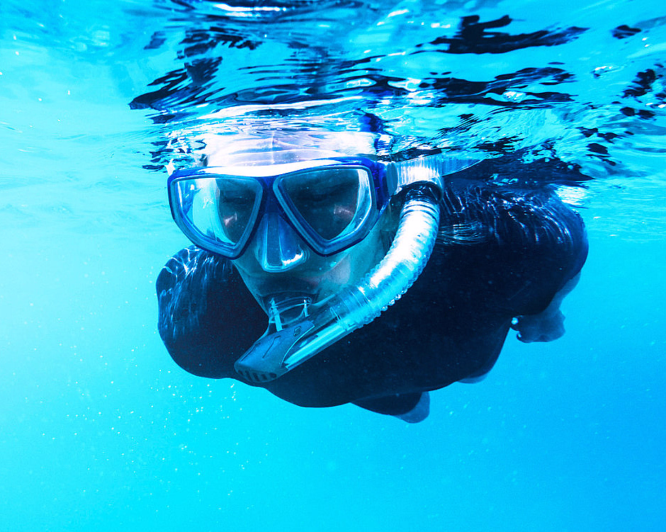 En person i svart våtdrakt og blå dykkermaske med snorkel svømmer i klart, blått vann med lysrefleksjoner på overflaten.