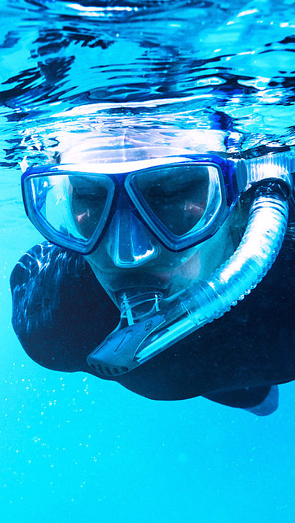 En person i svart våtdrakt og blå dykkermaske med snorkel svømmer i klart, blått vann med lysrefleksjoner på overflaten.