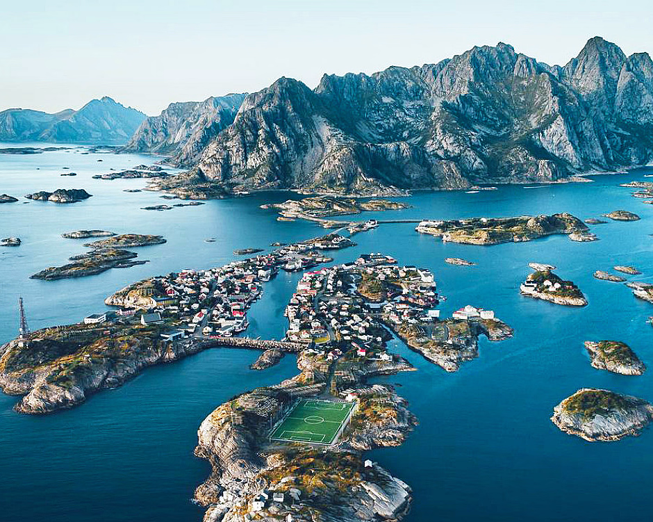 Flyfoto av en idyllisk kystlandsby med en grønn fotballbane på en liten øy, omgitt av blått hav og dramatiske fjell i bakgrunnen.