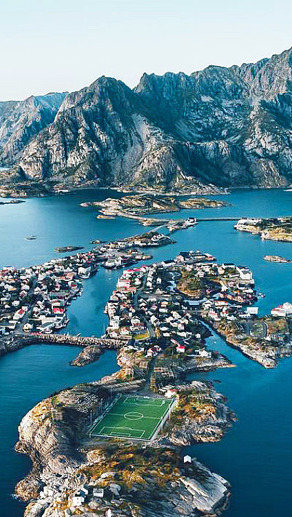 Flyfoto av en idyllisk kystlandsby med en grønn fotballbane på en liten øy, omgitt av blått hav og dramatiske fjell i bakgrunnen.