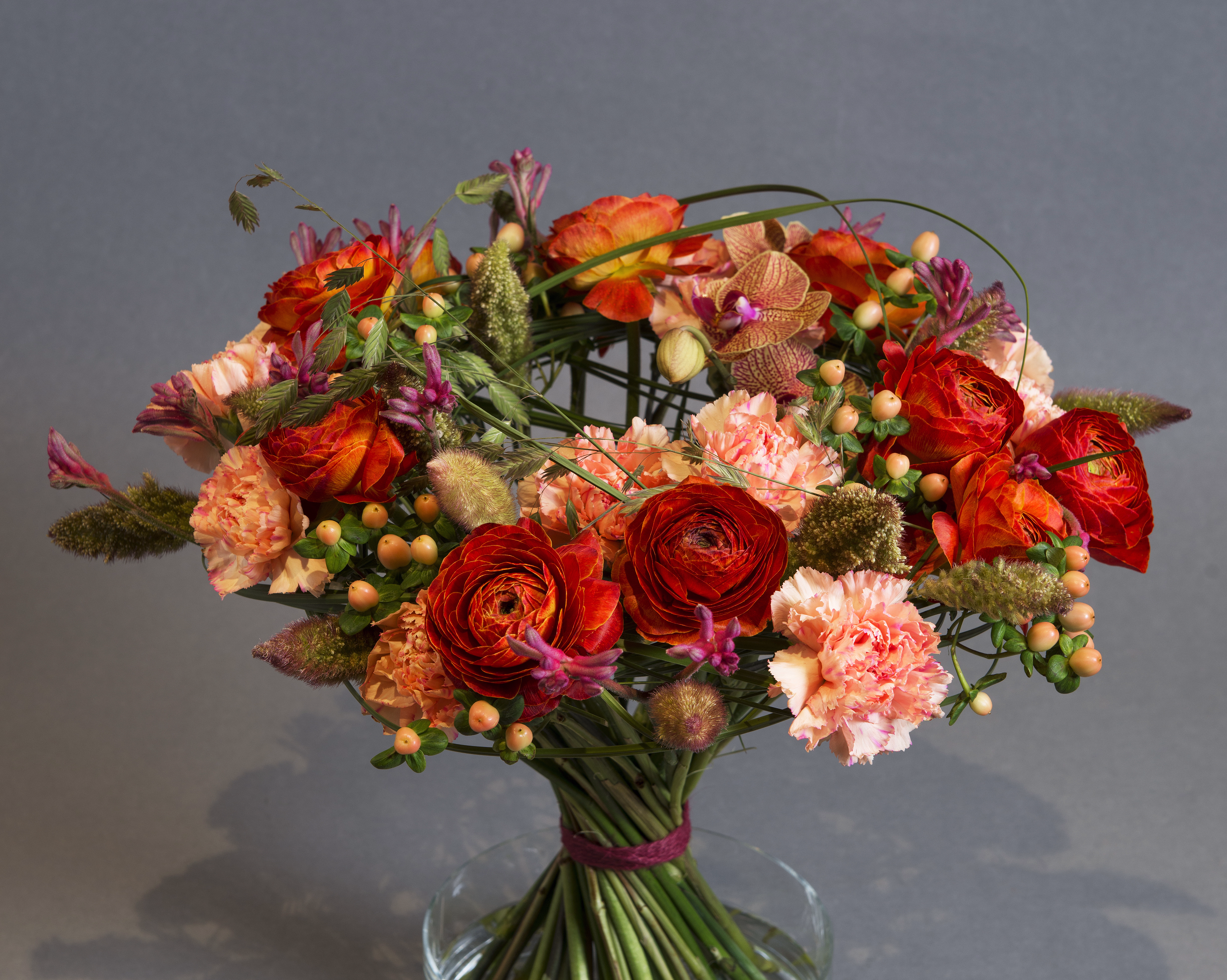 En fargerik blomsterbukett med oransje og røde roser, rosa nelliker og små bær i en klar glassvase mot en grå bakgrunn.