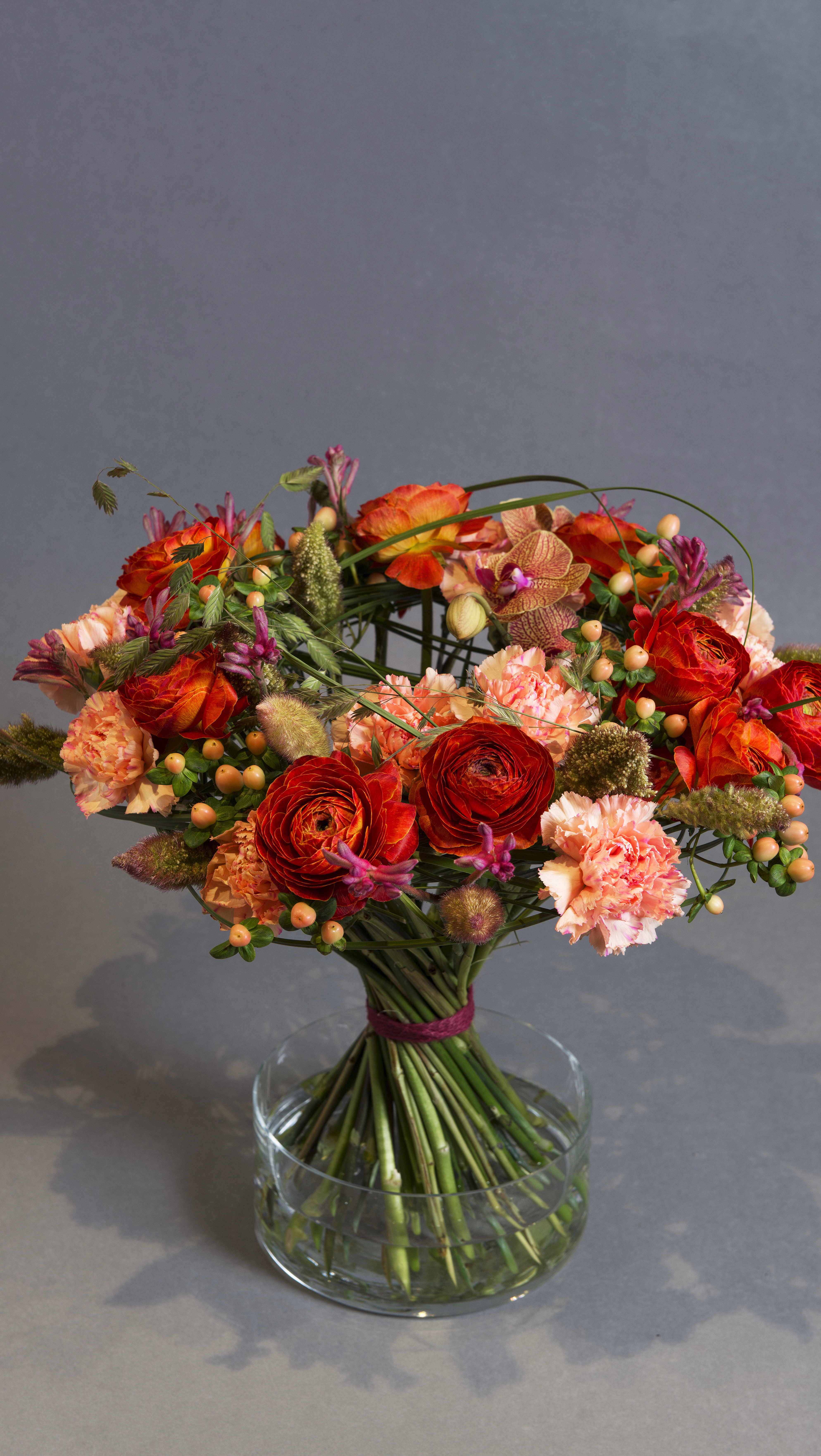 En fargerik blomsterbukett med oransje og røde roser, rosa nelliker og små bær i en klar glassvase mot en grå bakgrunn.