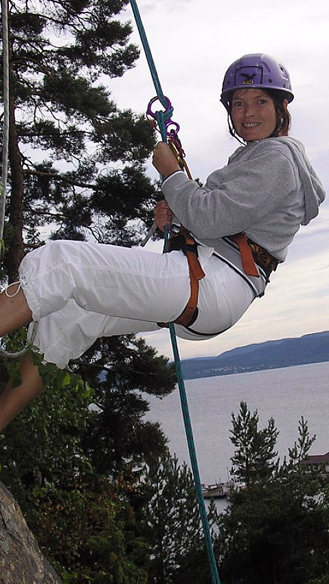 En person klatrer på en fjellvegg iført lilla hjelm og grå hettegenser, med utsikt over vann og åser i bakgrunnen.