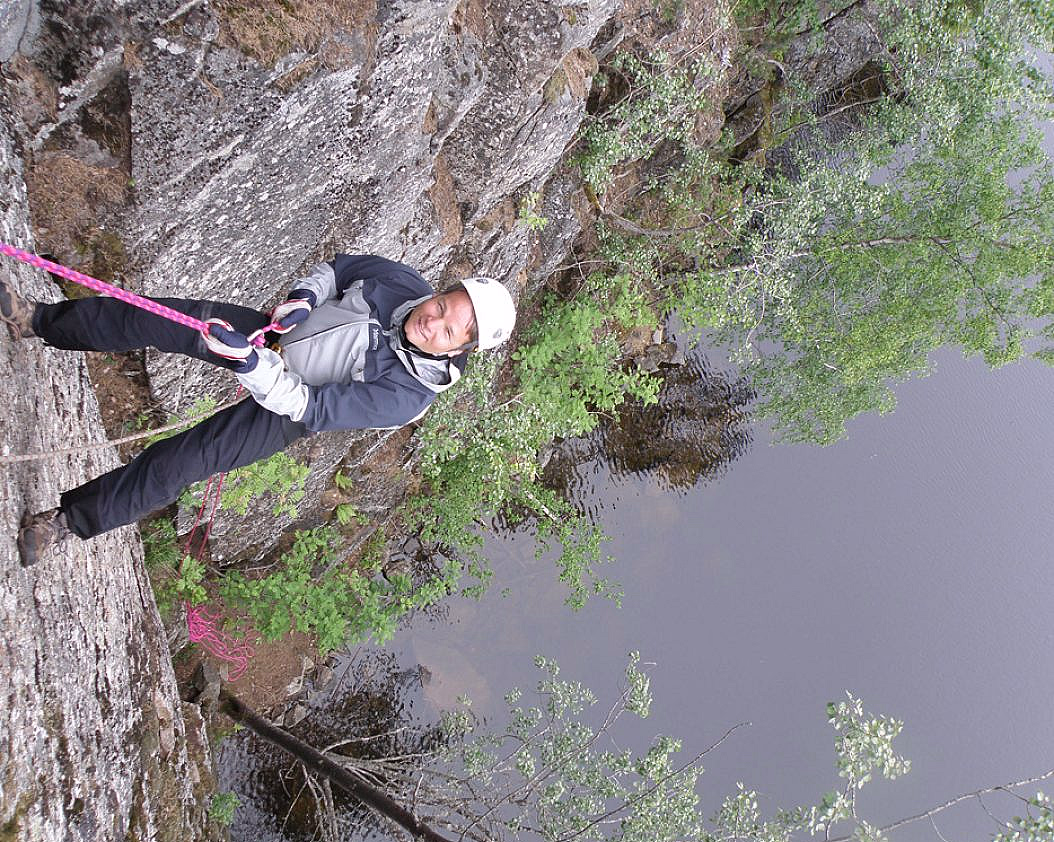 En person med hvit hjelm og grå jakke rappellerer ned en fjellvegg med et rosa tau, med vann og grønne trær i bakgrunnen.